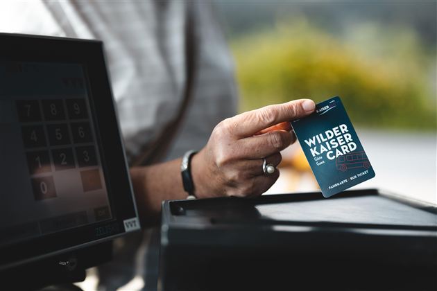 A person is holding a Wilder Kaiser Card in their hand. In the background, a cash register terminal is visible.