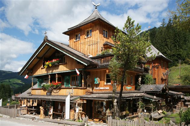 Ein traditionelles, holzverkleidetes Alpenhaus mit vielen bunten Blumen und einer malerischen Umgebung. Der Himmel ist blau mit einigen Wolken und grünem Wald im Hintergrund.