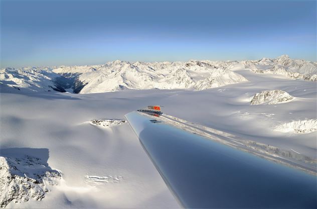 Eine beeindruckende Schneelandschaft mit majestätischen Bergen im Hintergrund. Im Vordergrund ist ein schimmernder, reflektierender Flügel eines Flugzeugs zu sehen.