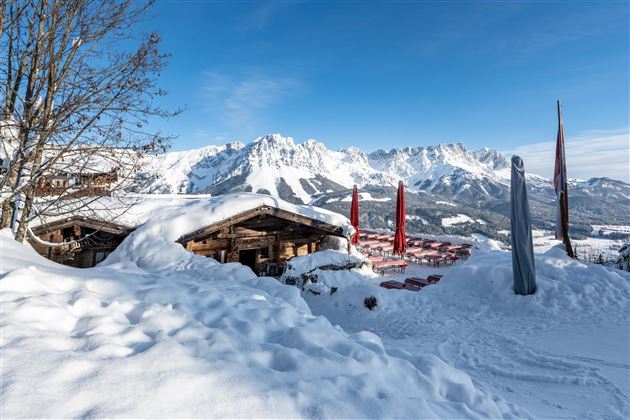 Eine winterliche Landschaft mit schneebedeckten Hütten und majestätischen Bergen im Hintergrund. Der Himmel ist klar und die Sonne scheint auf die schneebedeckte Fläche.