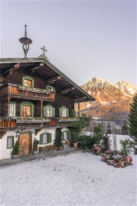 A traditional wooden house in the snow with balconies and flowers. Snow-covered mountains can be seen in the background.