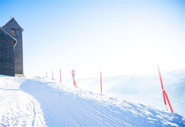 A snow-covered slope with red markings and a small church in the background. The landscape is bright and radiates a winter tranquility.