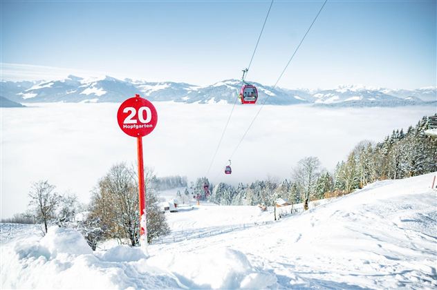 A snowy landscape with a ski lift. In the background, snow-covered mountains and a clear sky can be seen.