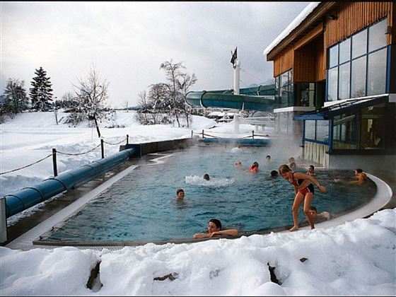 Een buitenzwembad in een winterse omgeving, omringd door sneeuw. Meerdere mensen genieten van het warme water en er is een glijbaan naast het bad.