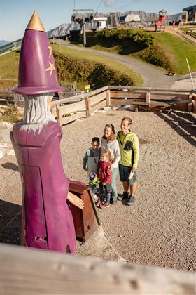 A group of children is standing in front of a colorful wizard figure outdoors. In the background, green meadows and some buildings can be seen.