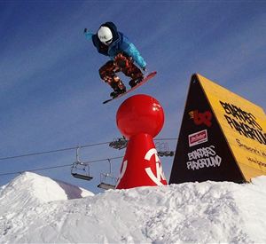 Een snowboarder springt over een rode koepel op een met sneeuw bedekte pistes. Op de achtergrond zijn skiliften en een reclamebord zichtbaar.
