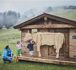 Een houten huis met een houten beeld van een koe staat op een weiland. Twee kinderen en een vrouw staan ervoor en verkennen het beeld.