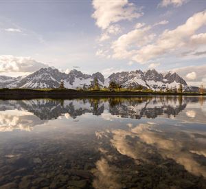 Eine malerische Berglandschaft mit schneebedeckten Gipfeln und einem ruhigen Wasser, das die Wolken spiegelt. Die Szenerie ist friedlich und einladend.