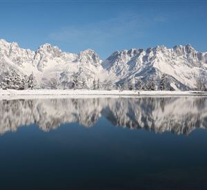 Eine beeindruckende Berglandschaft mit schneebedeckten Gipfeln und einem ruhigen See im Vordergrund. Der klare blaue Himmel spiegelt sich im Wasser.