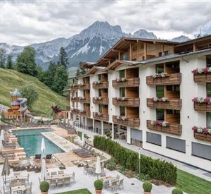 Een modern hotel met houten bekleding en balkonbloemen. Op de achtergrond zijn majestueuze bergen en een zwembad met ligstoelen te zien.