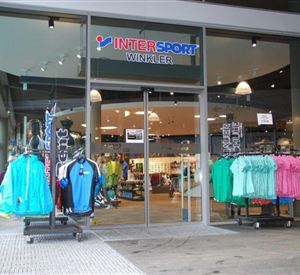 Een winkelfaçade met de naam "INTER SPORT WINKLER". Voor de deur staan verschillende kledingrekken met kleurrijke sportkleding.
