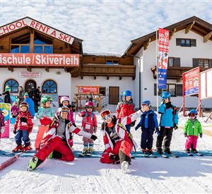 A ski school with a colorful building in the background. In front of the building, ski instructors and children in ski clothing are standing on snow.