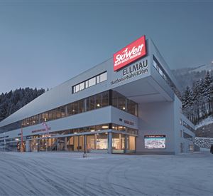 Een modern gebouw met grote ramen en een rood bord dat "Skisport Ellmau" toont. Het ligt in een besneeuwde omgeving, omringd door bomen en bergen.
