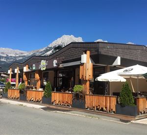 Ein gemütliches Restaurant mit einer Holzterrasse und Sonnenschirmen. Im Hintergrund sind majestätische Berge und ein blauer Himmel zu sehen.