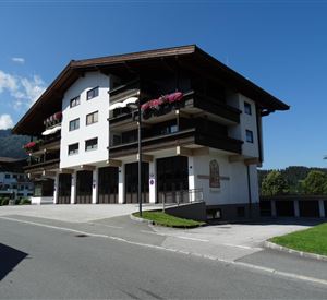 Een modern gebouw in alpine stijl met balkons en bloemen. De omgeving is groen en zonnig, met uitzicht op de bergen.
