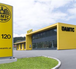 A modern building of the ÖAMTC with a striking yellow facade. In the foreground is a large sign with the inscription and the ÖAMTC website.