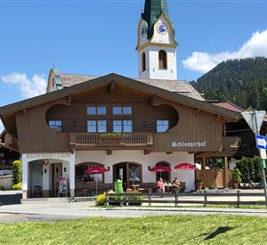 A traditional alpine inn with a characteristic clock tower. In the foreground, there are tables with red umbrellas and a green meadow.