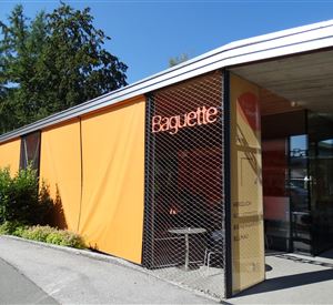 A modern building with a striking orange facade and the inscription "Baguette". Surrounded by green plants and under a clear blue sky.