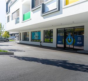 Een modern gebouw met grote ramen en een zakelijke gevel. Op de voorgrond zijn de parkeerplaats en een auto zichtbaar.