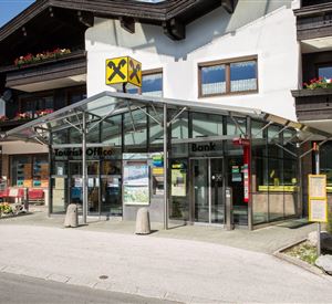 Eine moderne Bank mit einem großen Glasvordach. Sie befindet sich an einer belebten Straße und ist von gepflegten Pflanzen umgeben.
