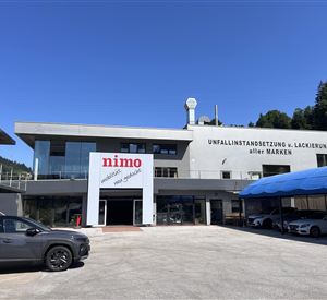 Een moderne autodealer met een grote glazen gevel en een heldere blauwe lucht. De locatie biedt diensten aan voor schadereparaties en spuitwerk.