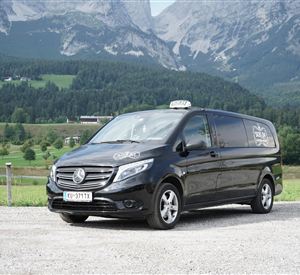 Ein schwarzer Van von Mercedes-Benz steht auf einer gravelartigen Fläche vor einer malerischen Berglandschaft. Im Hintergrund sind grüne Wiesen und hohe Berge sichtbar.