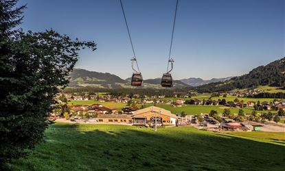 Eine Seilbahn, die über eine grüne Wiese schwebt, mit Blick auf ein kleines Dorf und die umliegenden Berge. Der Himmel ist klar und blau.