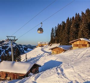 Ein malerisches Winterland mit schneebedeckten Hütten und einer Seilbahn. Hohe Tannenbäume umgeben die Szene, während die Sonne am klaren Himmel scheint.