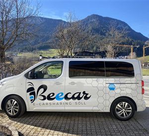 Een E-Carsharing-voertuig van het merk Beecar staat aan de rand van de straat. Op de achtergrond zijn bergen en een blauwe lucht zichtbaar.