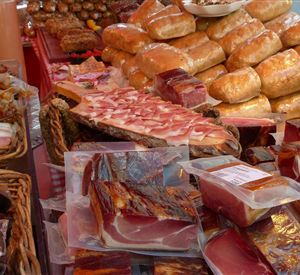 Een marktkraam met verschillende vleessoorten, waaronder ham en worst. Op de achtergrond zijn verse broodjes zichtbaar.