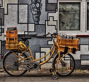 Ein gelbes Fahrrad mit zwei Kisten ist an einer Wand mit grafischer Gestaltung abgestellt. Im Hintergrund ist ein Fenster zu erkennen.