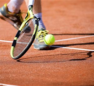 Een tennisbal stuitert op de zandbaan. Op de voorgrond is een tennisracket en een speler te zien.