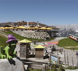 Een berglandschap met een restaurant en zonnige terrassen. Op de voorgrond staat een groene figuur met een paarse hoed en speeltoestellen voor kinderen.