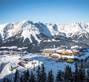 A wintry mountain landscape with snow-covered peaks and a ski resort. In the foreground, ski lifts and a mountain hut with seating are visible.