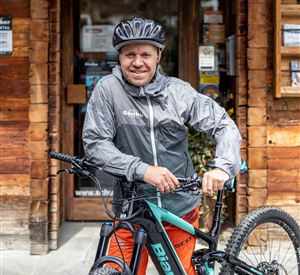 A man in a gray rain jacket stands next to a mountain bike. In the background, a wooden cabin is visible.