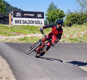 A mountain biker is riding on a winding track. In the background, the sign "Bike Saloon Söll" is visible.