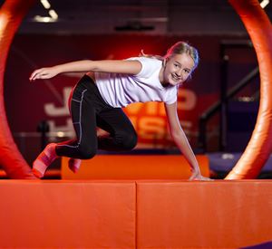 Een meisje springt over een oranje platform in een trampolinepark. De achtergrond is kleurrijk en uitnodigend ingericht.