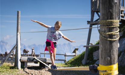 Een meisje balanceert op een touw in de bergen. Op de achtergrond zijn een mooi landschap en een houten structuur zichtbaar.