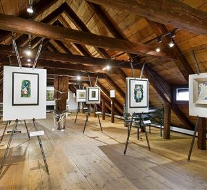 Een verbouwde zolder met houten plafonds. Er zijn verschillende kunstwerken in lijsten op standaards tentoongesteld.