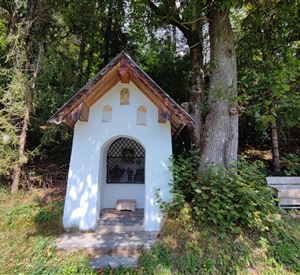 Een kleine kapel staat in een bosrijke omgeving. Naast de kapel staat een houten bank.