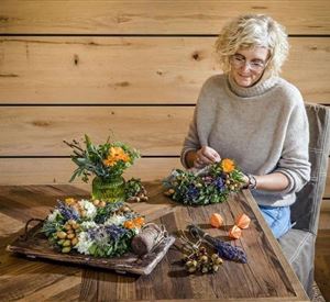 Een vrouw werkt aan een tafel met verschillende bloemenarrangementen. Op de achtergrond zijn houten oppervlakken zichtbaar, wat een gezellige sfeer creëert.