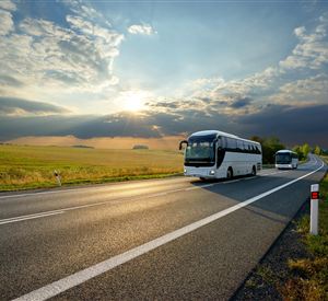 Twee touringcars rijden op een landelijke weg door een groene landschap. De lucht is bewolkt en de zon schijnt aan de horizon.