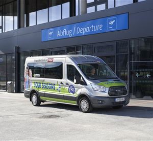 Ein Linienbus steht vor einem modernen Flughafengebäude. Über dem Bus ist das Schild "Abflug / Departure" sichtbar.