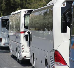 Een reeks witte toerbussen parkeert langs een straat. De omgeving is groen en zonnig.