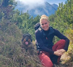 Een vrouw zit op een wandelpad naast haar hond. Ze is omringd door groene bomen en bergen.