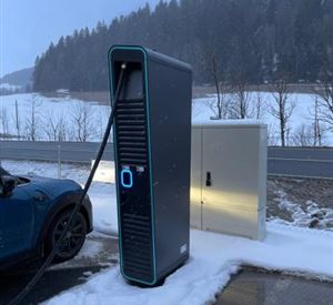 Een laadstation voor elektrische auto's staat op een met sneeuw bedekte parkeerplaats. Op de achtergrond zijn bomen en een winterlandschap te zien.