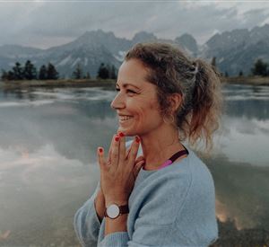 Een vrouw staat aan de oever van een rustig meer en lacht. Op de achtergrond zijn bergen en een bewolkte lucht te zien.