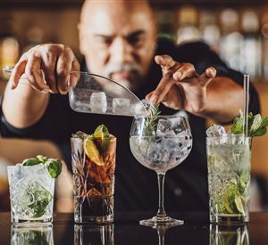 Een bartender mixt verschillende cocktails aan een bar. Op de voorgrond staan verschillende stijlvolle glazen met verse ingrediënten en ijs.
