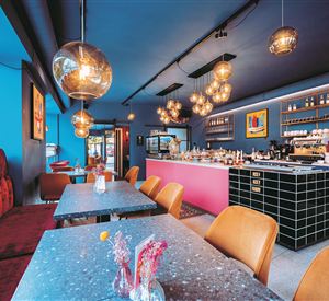 Een modern café met een stijlvolle blauwe wanddecoratie. De inrichting omvat gezellige tafels en kleurrijke verlichting.