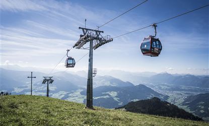 Eine Seilbahn fährt über eine grüne Wiese mit Bergen im Hintergrund. Der Himmel ist klar und blau.